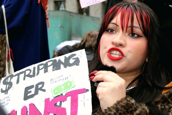 Stripper Alma Adore holder en plakat der det står "Stripping er Kunst". Hun er midt i en setning og er derfor hard i fjeset og ser sint ut.