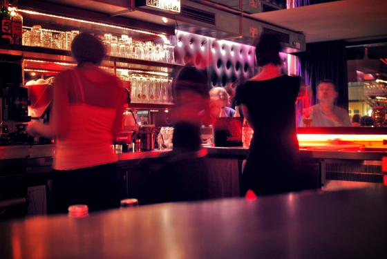 Rosa lys i en bar. Noen bartendere st&aring;r og shaker drinker, mens kundene venter. 