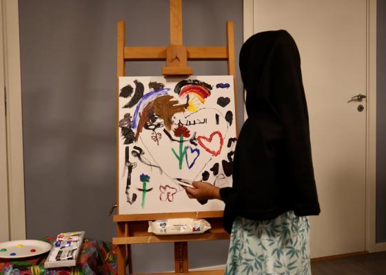Liten jente med hijab som maler p&aring; et lerret. Det er blomster, hjerter og arabisk skrift p&aring; maleriet. Hun st&aring;r med ansiktet mot maleriet. 