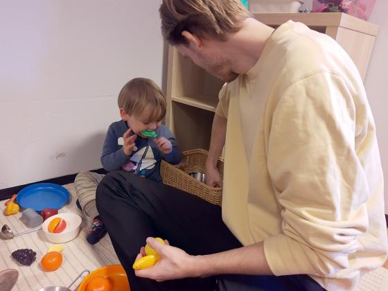 Barnehagel&aelig;rer Carl sitter p&aring; gulvet ved siden av en gutt som har en lekesalat i munnen.