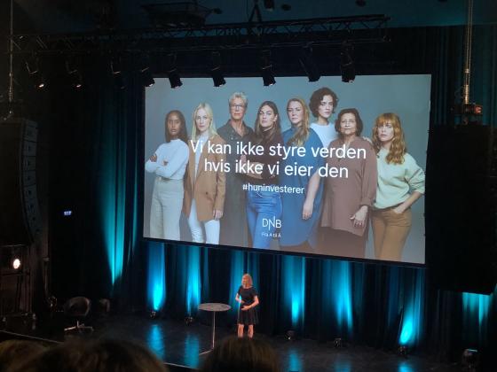 Aina Lemoen Lunde foran et stor publikum p&aring; Sentrum Scene i Oslo, med DNBs reklame for #HunInvesterer p&aring; lerret bak seg.