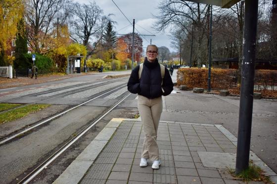 Blond kvinne med svart jakke, beige ryggsekk og m&oslash;rke briller st&aring;r og venter p&aring; trikken utenfor Universitetet i Oslo, avd. Blindern