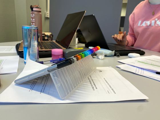 Bilde av et rotete bord med to pcer, b&oslash;ker, tusjer og energidrikk. To jenter som sitter og jobber med eksamen