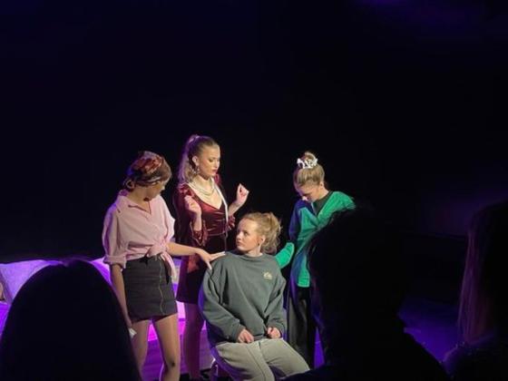 P&aring; en liten teaterscene st&aring;r ei jente i posete kl&aelig;r. Rundt henne st&aring;r fullt sminkede jenter i trange kl&aelig;r og trykker seg p&aring; henne. 