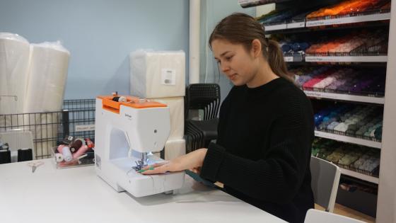 En jente bruker symaskin, med en stor hylle med garnn&oslash;ster bak seg. 