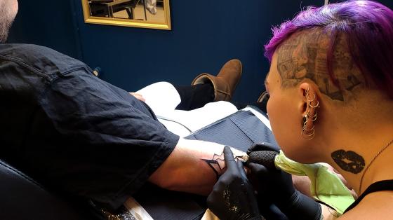 Ei dame tatoverar eit lyn p&aring; armen til ein mann. Tatov&oslash;ren har lilla h&aring;r.