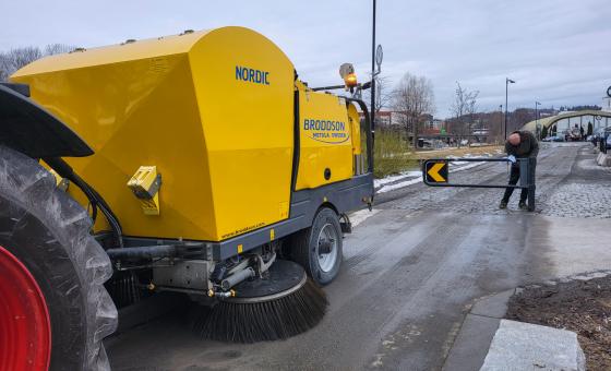 Bilde av hengeren p&aring; en feiebil, som er stor og gul. I bakgrunnen er det en mann som lukker en bom.