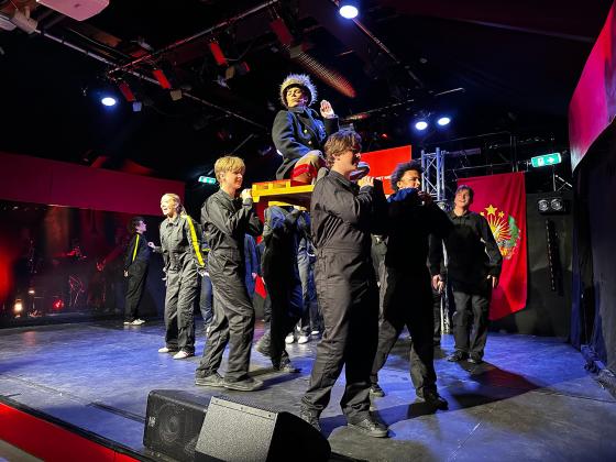 Akt&oslash;rer i svarte drakter g&aring;r p&aring; en scene og b&aelig;rer en annen akt&oslash;r p&aring; en b&aring;re. 