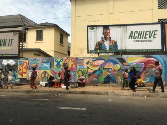 Bildet viser et fortau i Ghana. Det g&aring;r flere personer p&aring; fortauet, og bak dem er en vegg dekket i fargerik graffiti. 