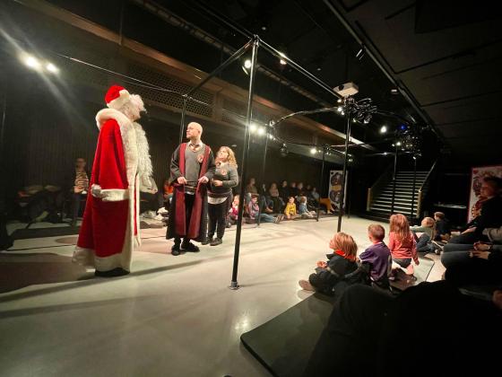 To skuespillere utkledt som Harry Potter og Hermine i et lite lyssatt scenerom. De ser p&aring; en mann i julenissedrakt. I enden av rommet er en hvitkledd trapp. I taket er det lys. Langs sidene sitter det barn p&aring; matter. 