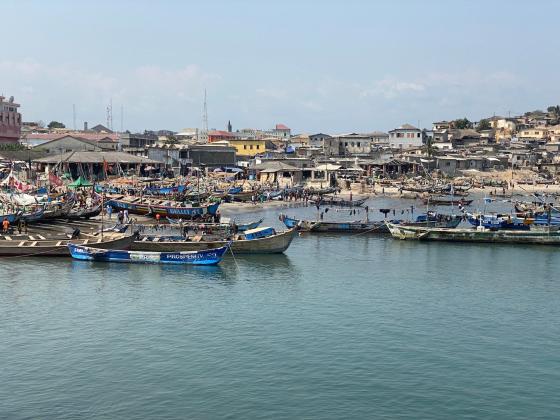 vi ser et bl&aring;tt vann med b&aring;ter, en strand og flere hus i bakgrunnen