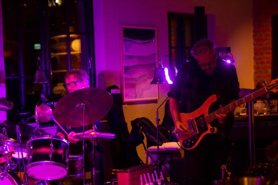 En mann med svarte briller sitter bak et trommesett. En mann med gr&aring;tt h&aring;r spiller bass. 