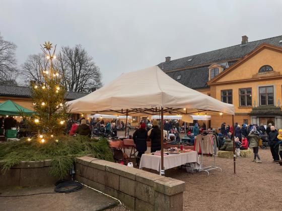 Bildet viser julemarkedet p&aring; Bogstad G&aring;rd. Man kan se et juletre med lys, et hvitt telt som holder regnet unna noen som selger produkter, og mange folk i bakgrunnen. 