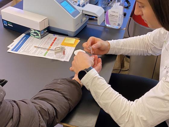 Bildet viser en kvinne som f&aring;r sjekket blodsukkeret sitt. Hun har blitt stukket i fingeren, s&aring; det er litt blod p&aring; fingeren. Frisklivsveilederen holder et utstyr til &aring; hente noen dr&aring;per til m&aring;lingen.