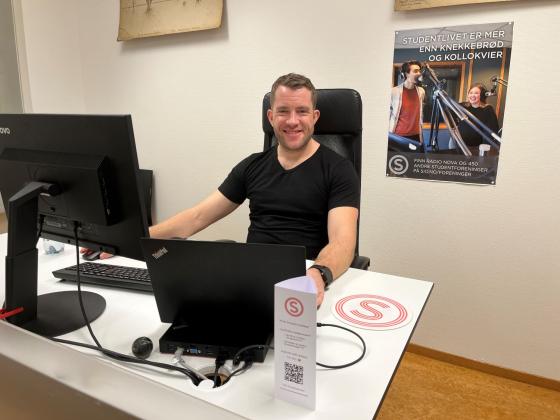 Bildet viser en mann i svart T-skjorte som smiler til kameraet. Han sitter p&aring; et kontor, med to PC skjermer foran seg. 