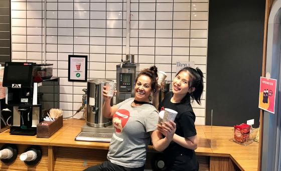 To kvinner st&aring;r i kafeterian p&aring; Oslo Met og poserer med kaffe foran kaffehj&oslash;rnet.