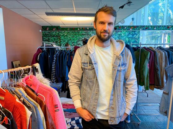 Mannen st&aring;r foran noen kl&aelig;rstativer med hendene i lomma. Han har p&aring; seg en hvit tskjorte med en denim jeansjakke over.