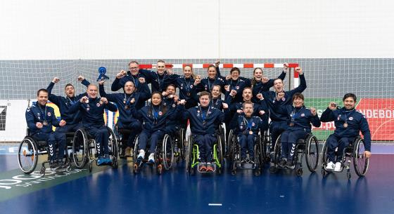 Alle spillerne p&aring; rullestolh&aring;ndballandslaget og st&oslash;tteapparatet bak st&aring;r foran et m&aring;l og jubler med bronsemedaljen rundt halsen