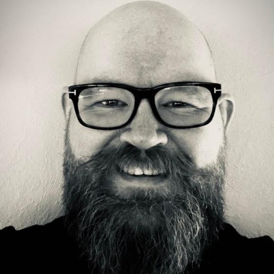 Gr&aring;tt og hvitt portrettbilde av Lars Martin Torp. Med stort smil, skallet hode, svarte briller og stort skjegg