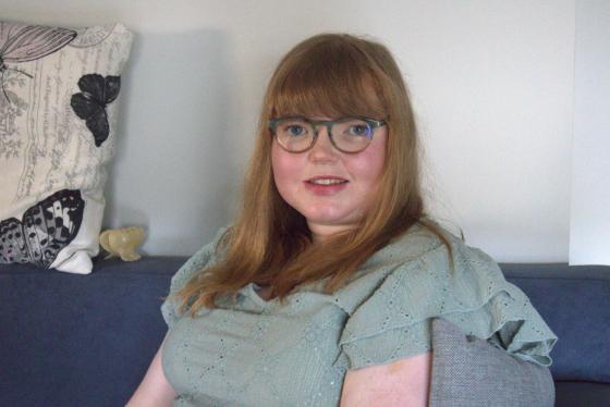 Kvinne p&aring; 31 &aring;r med r&oslash;dt h&aring;r og briller sitter i gr&aring; sofa og ser p&aring; kameraet.