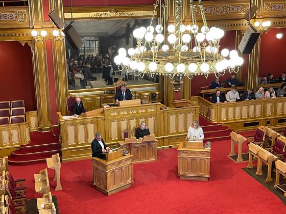 P&aring; bildet ser man en stor sal, med gullmalt interi&oslash;r, og r&oslash;de tepper kler gulvet. Vi ser et stort kunstverk, en stor lysekrone. Vi ser 16 mennesker p&aring; bildet. En kvinne har p&aring; seg hvite kl&aelig;r.