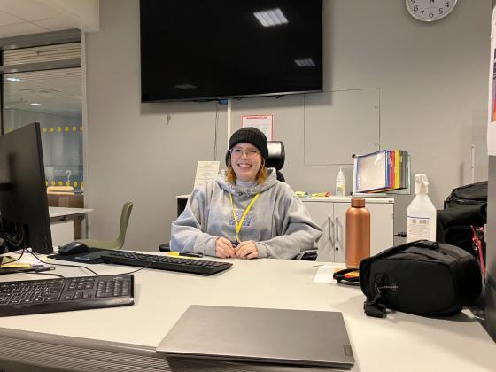 Dame (Andrea Hansen) som sitter bak en pult inne p&aring; et bibliotek. Foran henne p&aring; bordet st&aring;r skjermen til en stasjon&aelig;r pc og et tastatur, pluss en laptop, en kamerabag, drikkeflaske, antibac.