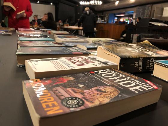 mange b&oslash;ker ligger p&aring; et bord. Personer st&aring;r i bakgrunnen