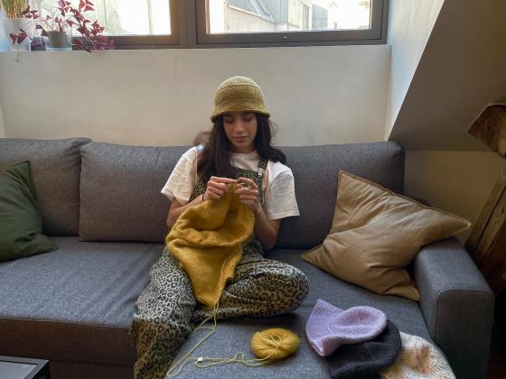 Nesrin Alma Kalayci sitter i sofaen og strikker p&aring; en gul genser. 