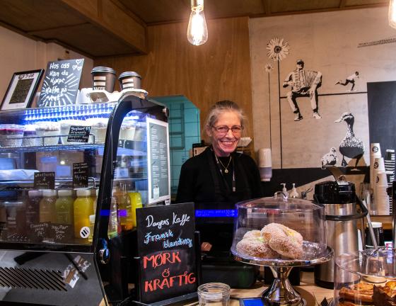 En gr&aring;h&aring;ret dame med briller smiler mot kamera, fra bak et kasseapparat. Foran henne st&aring;r et fat med skoleboller. 