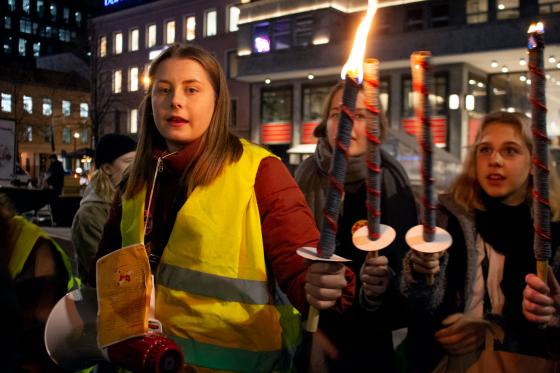 Jente ser i kamera mens hun holder en opptent fakkel. Hun tenner faklene til to andre ogs&aring;.