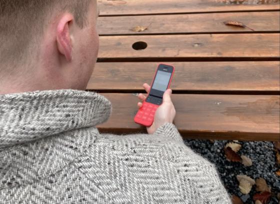 Bilde av en mann med gr&aring; ullk&aring;pe som sitter p&aring; en benk. Han holder en r&oslash;d Nokia - knappetelefon. 