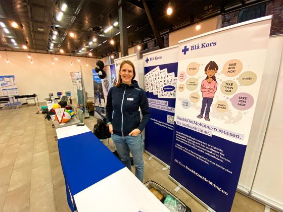 En kvinne i bl&aring; genser st&aring;r foran et bord. 