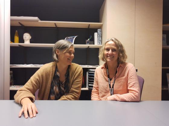 To kvinner som sitter. Den ene ser p&aring; den andre, mens den andre ser i kamera.