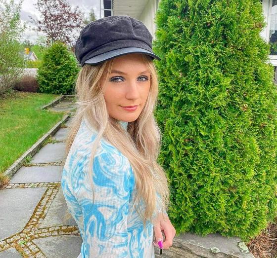 Sanna har p&aring; seg en svart lue og st&aring;r foran noen busker. Hun har p&aring; seg en lysebl&aring; genser med hvite b&oslash;lger. Hun har langt, blondt h&aring;r og smiler til kamera. 