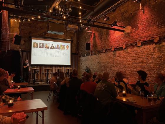 En stor scene hvor programlederes st&aring;r med en skjerm bak seg. Publikum ser opp mot skjermen og klapper. 