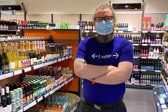 Butikksjef, Tom Erik Wiklund, st&aring;r foran hyllene p&aring; Orkanger Vinmonopolet med munnbind og t-skjorte, som oppfordrer kunder til &aring; holde 1 meter avstand.