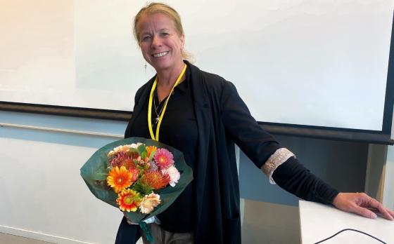 Her er Tonje, ei dame som st&aring;r foran en prosjekter med en blomsterbukett i h&aring;nda. Hun har p&aring; seg svarte kl&aelig;r, og har h&aring;ret i hestehale. 