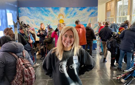 En smilende, ung dame st&aring;r  foran et bord, innend&oslash;rs. Det er mange mennesker som har ski i hendene i samme rom.
