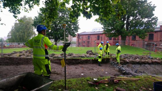 En gruppe mennesker st&aring;r i bakgrunnen med verneutstyr og snakker. En dame st&aring;r i forgrunnen med en type m&aring;leinstrument og har ogs&aring; p&aring; seg verneutstyr.