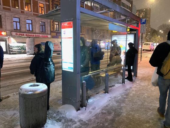 Bildet viser flere mennesker som venter p&aring; bussen p&aring; T&oslash;yengata buss-stopp, vendt mot Super Bawan Dagligvare. Det er litt m&oslash;rkt ute, og sn&oslash;er faller ned. Det er sn&oslash; p&aring; bakken. Personene st&aring;r med ryggen til fotograf.