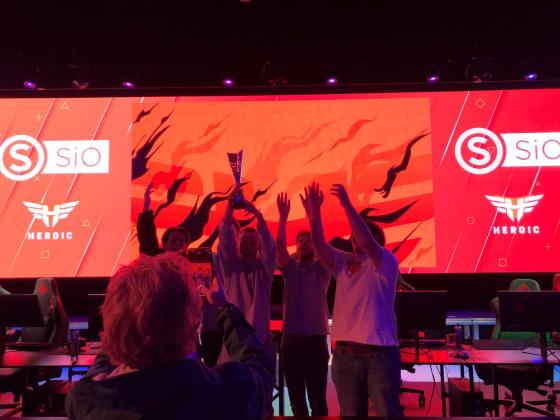 Fem jublende personer st&aring;r foran p&aring; en scene og l&oslash;fter en pokal. 