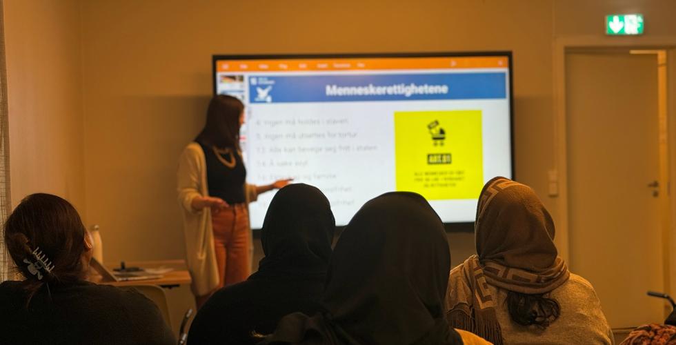 Seminar. Fem personen som sitter foran en skjerm. Foredragsholder st&aring;r ved siden av skjermen og peker.