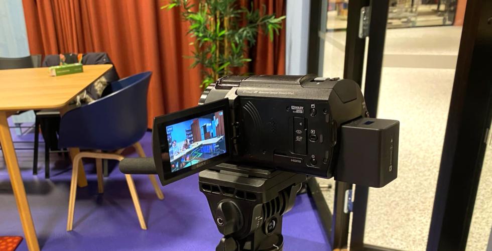 Et filmkamera som filmer to personer, som sitter og snakker p&aring; et fargerikt m&oslash;terom.