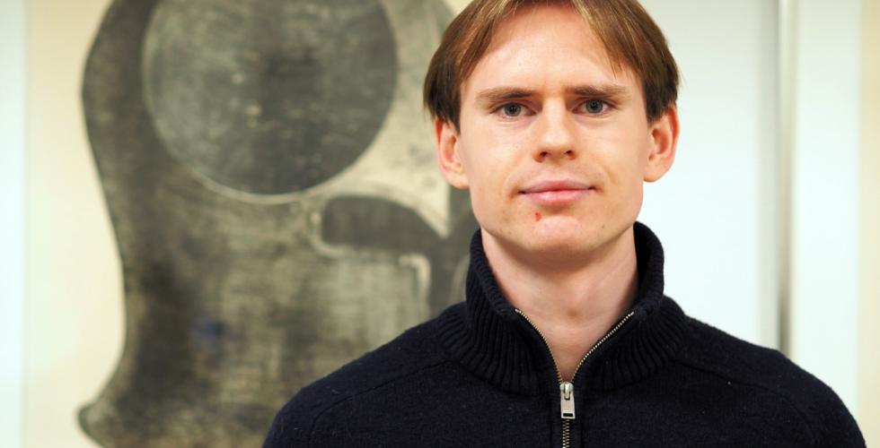 Mann st&aring;r foran et svart maleri, han har p&aring; seg en m&oslash;rkebl&aring; genser.