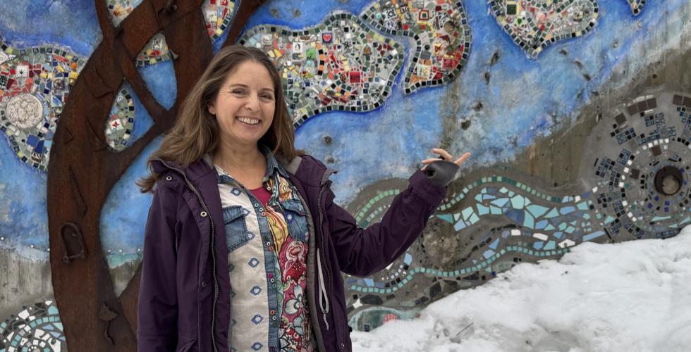 Jannicke Wiel st&aring;r foran en vegg dekorert med fargerike steiner i m&oslash;nster av et tre. Smiler og l&oslash;fter armen for &aring; vise frem. 