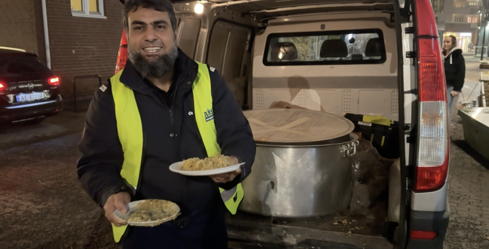 En mann i refleksvest er glad til &aring; servere to retter. Han st&aring;r foran en bil med &aring;pent bagasjerom. I bagasjerommet st&aring;r en stor gryte med mat. 