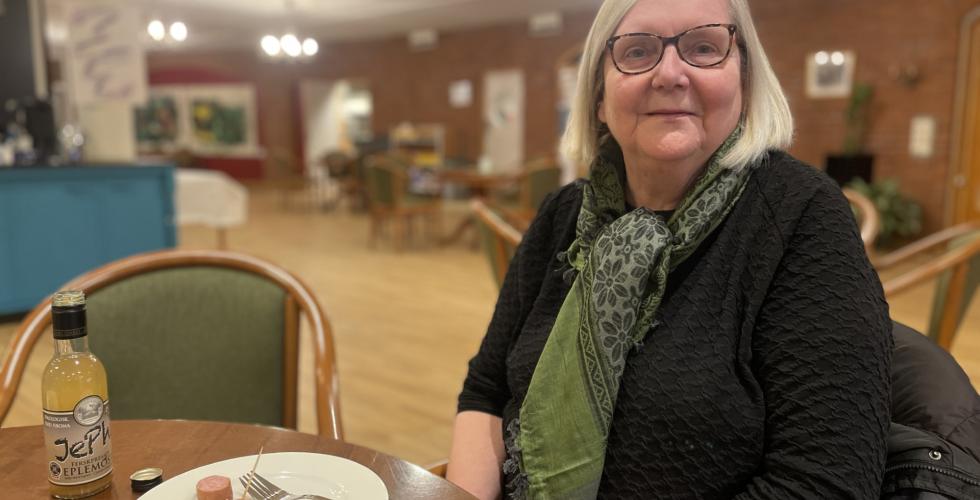Solveig sitter ved et bord og spiser p&oslash;lse og drikker eplemost. 