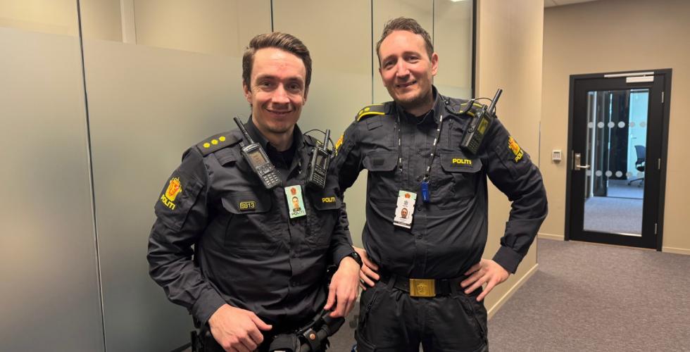 To politibetjenter som smiler inne p&aring; politistasjonen oppstilt ved siden av hverandre. 