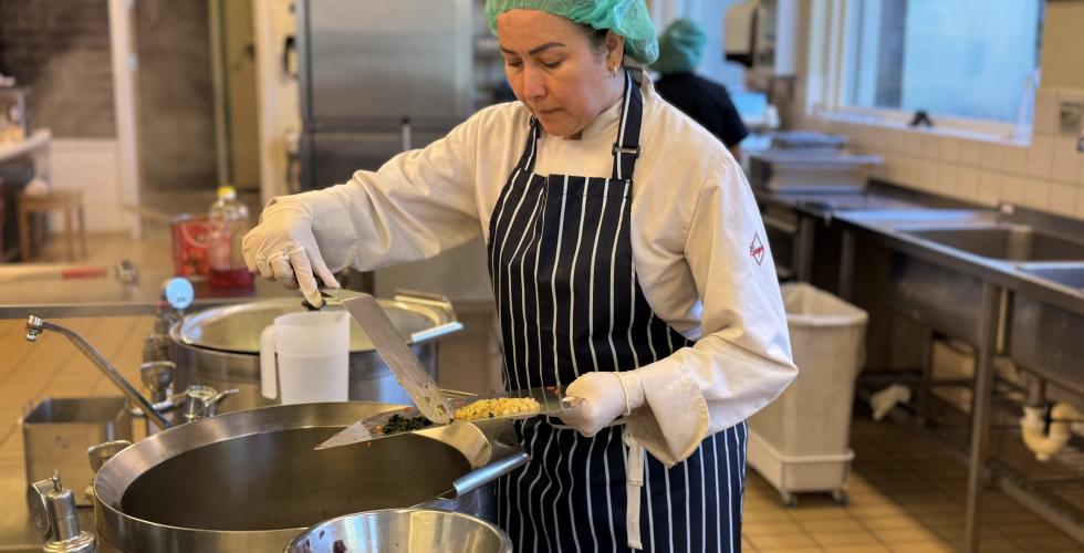 Andrea st&aring;r p&aring; et kj&oslash;kkenet ovenfor en kjele og skyver gr&oslash;nnsaker fra en fj&oslash;l over i kjelen. Hun har p&aring; forkle og h&aring;rnett og konsentrerer seg p&aring; matlagingen. 