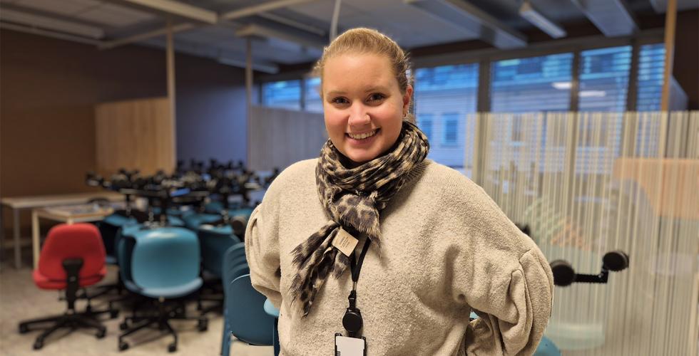 En dame står foran et rom under oppussing fylt med stoler. Hun har håret opp i en hestehale, og skjerf og genser på. Hun smiler og har hendene bak ryggen.